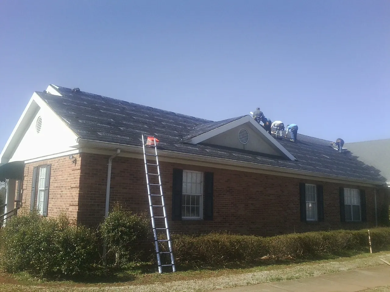 Crew tearing shingles off a brick ranch roof with a ladder set — Metal Roof Repair work by our crew in Oyens