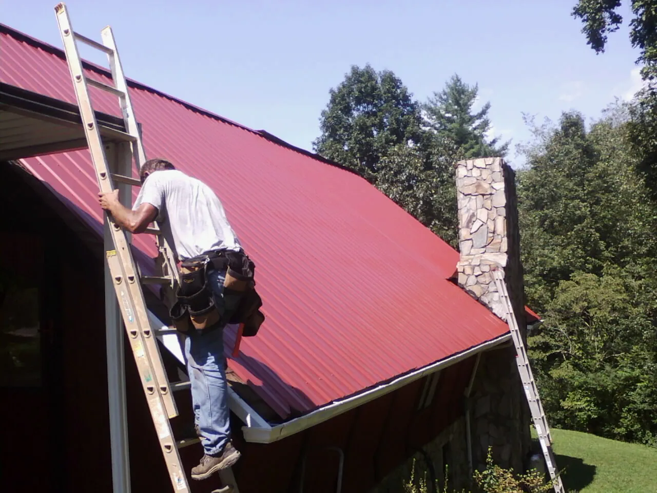Roofer on a ladder beside a red metal roof and stone chimney — Soffit & Fascia Repair by Roofing Brothers in Oyens